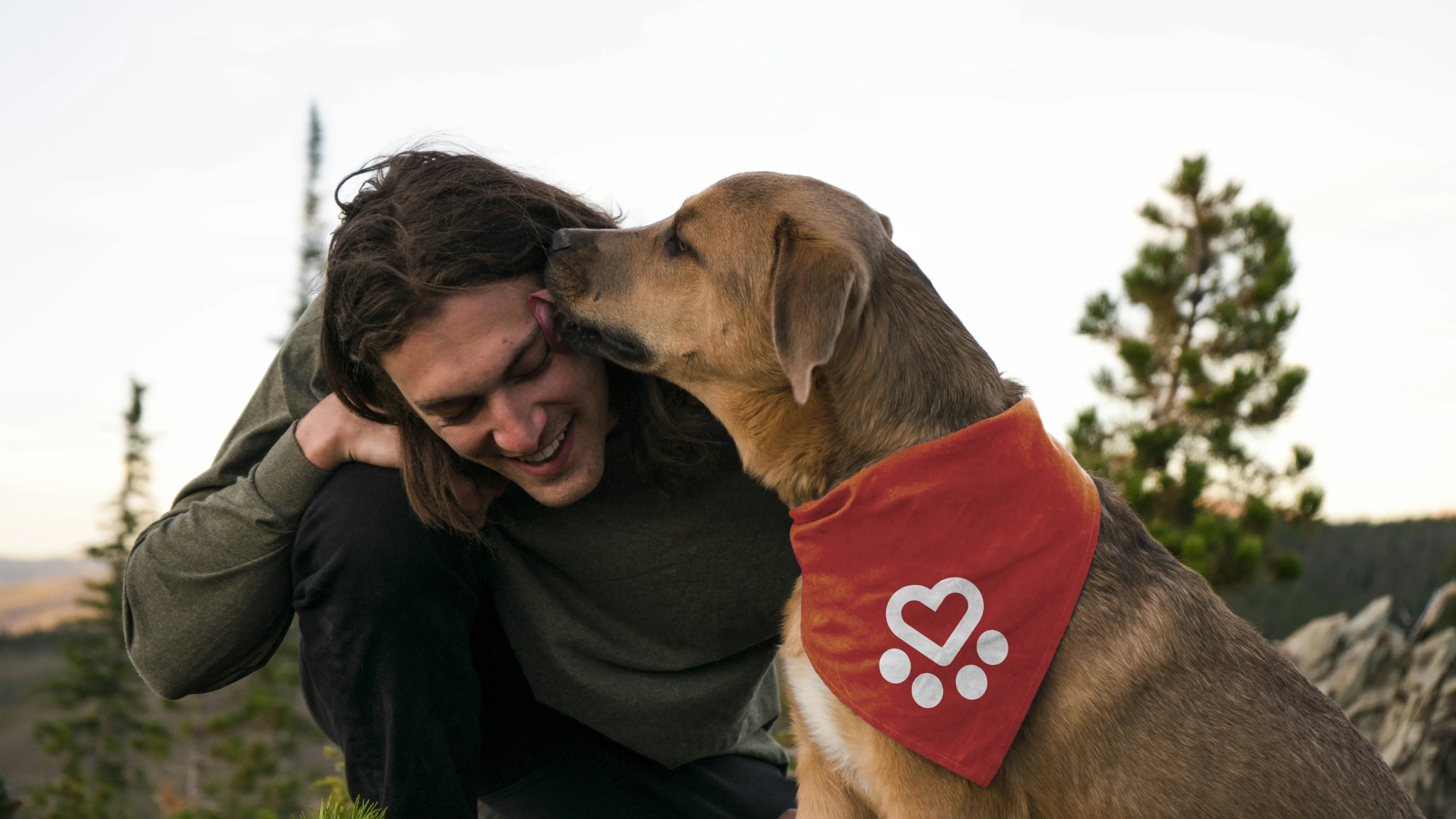 A dog rescue pup in a red bandana with a heart and paw design licks a smiling person's face outdoors near trees.