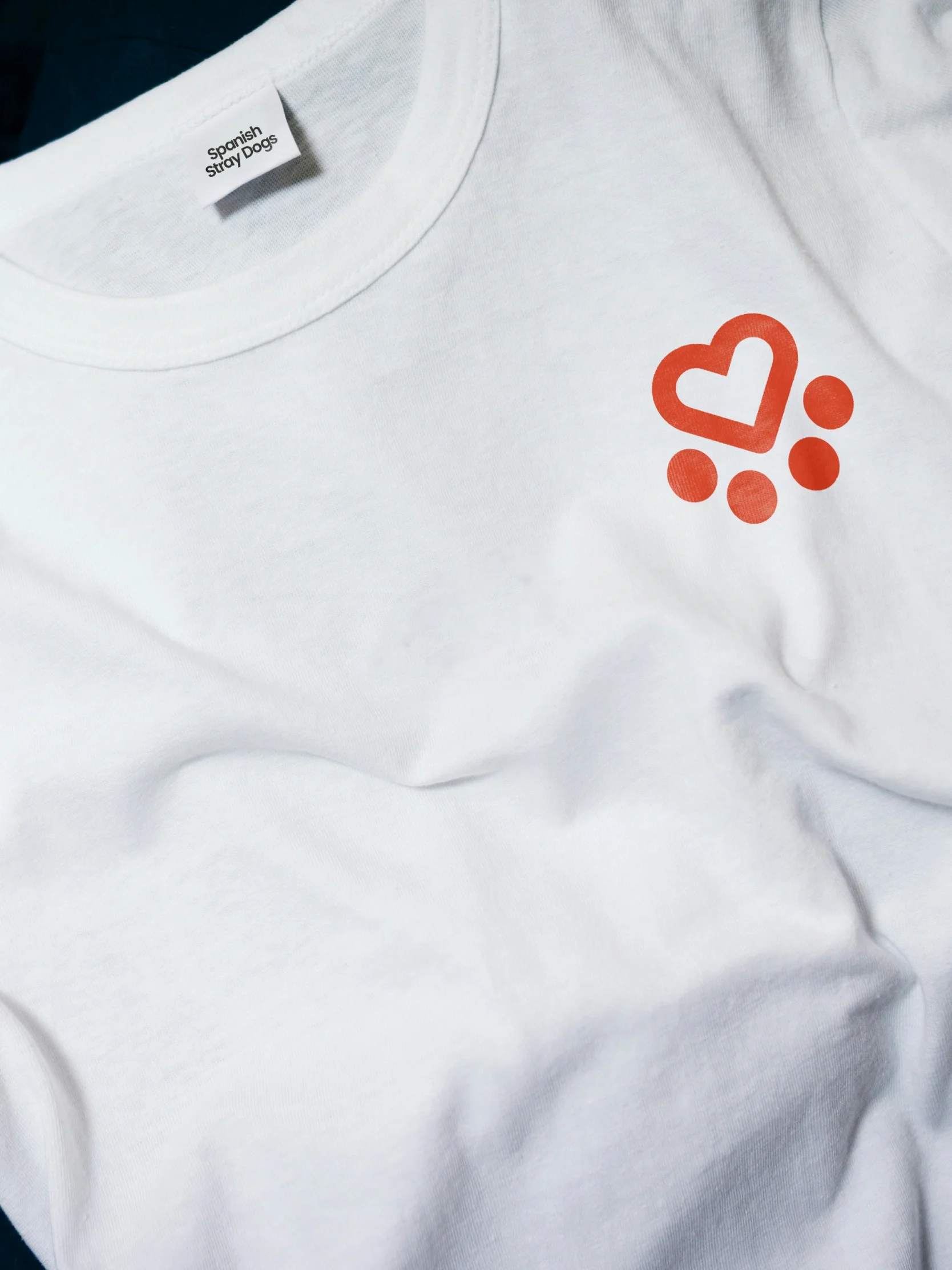 A white charity t-shirt featuring a small red heart and dot, supporting Spanish Stray Dogs, with a visible Stanley/Stella neck tag.