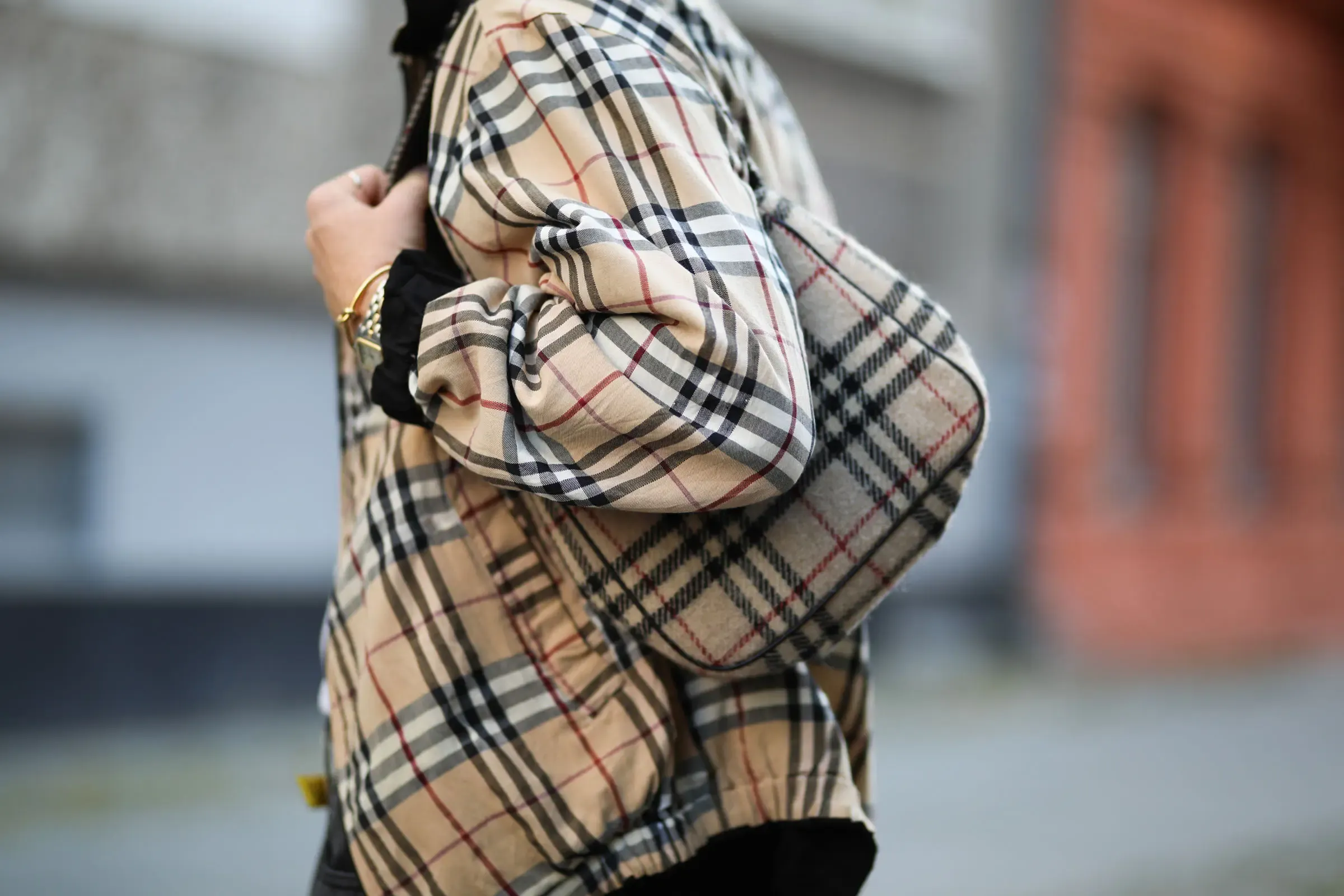 Close-up of a person on a blurred city street wearing an oversized jacket in the beige, black, white, and red Burberry check, one hand gripping the shoulder strap of a matching checked bag. The pattern is unmistakable even though no logo and no full garment is visible.