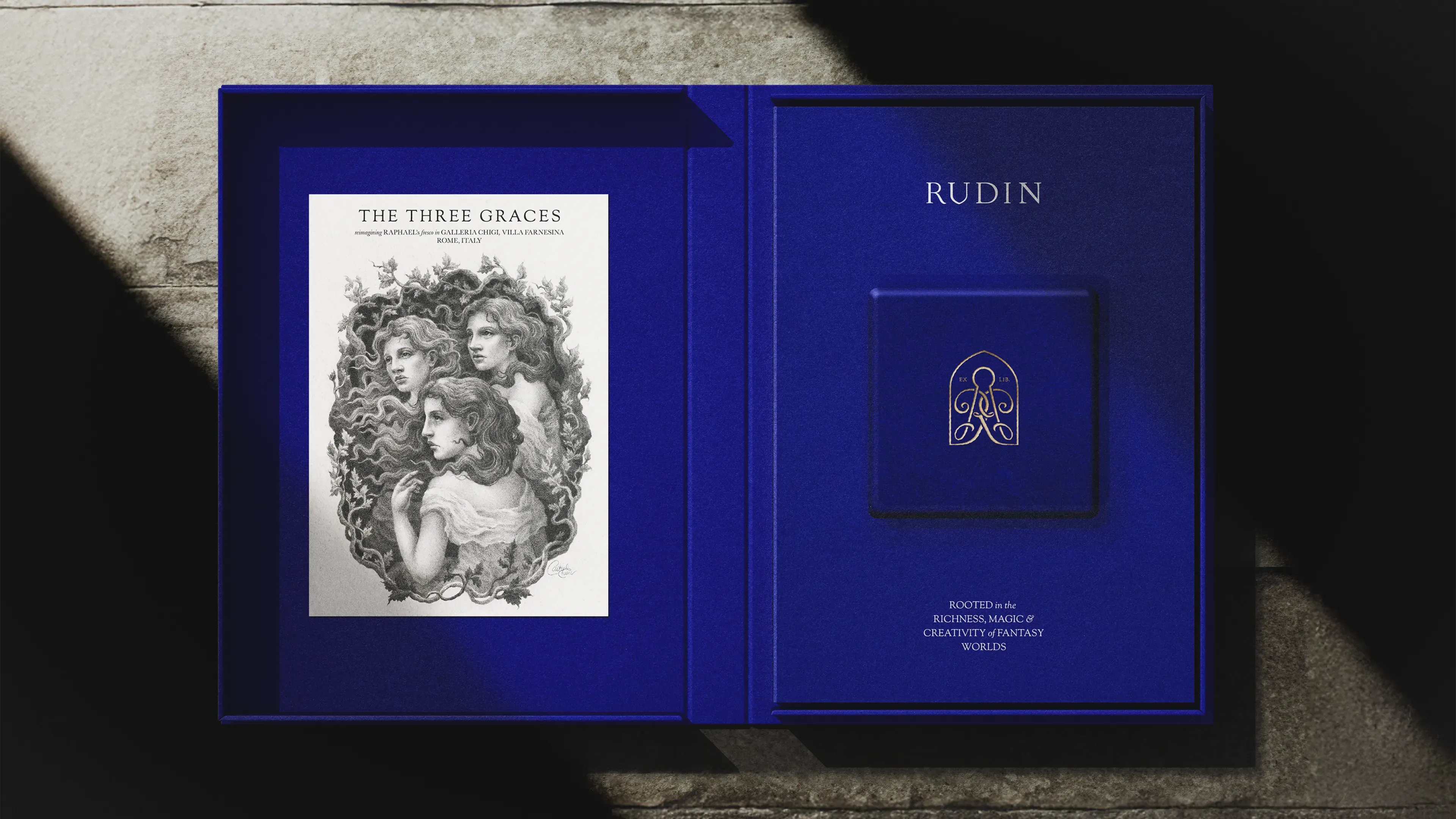 An open Rudin jewellery box revealing a deep royal blue interior. On the left side sits a printed illustration card titled The Three Graces, depicting three women with flowing hair in a pencil-drawn oval. On the right, a blue panel holds the embossed Rudin logo above the words Rooted in the richness, magic of creativity and fantasy worlds