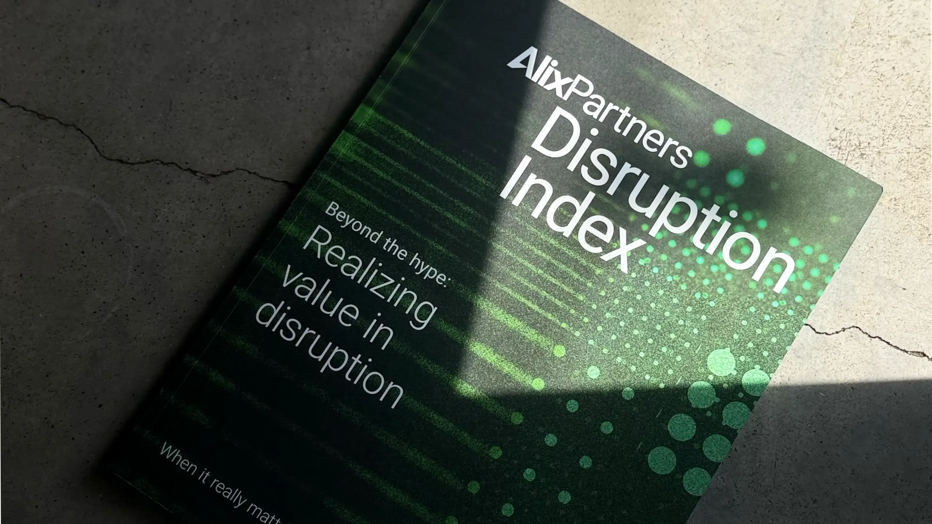The printed AlixPartners Disruption Index resting on a concrete floor in raking light. The dark cover carries the title in white, with the strapline 'Beyond the hype: realizing value in disruption', over green scan-line and particle imagery.