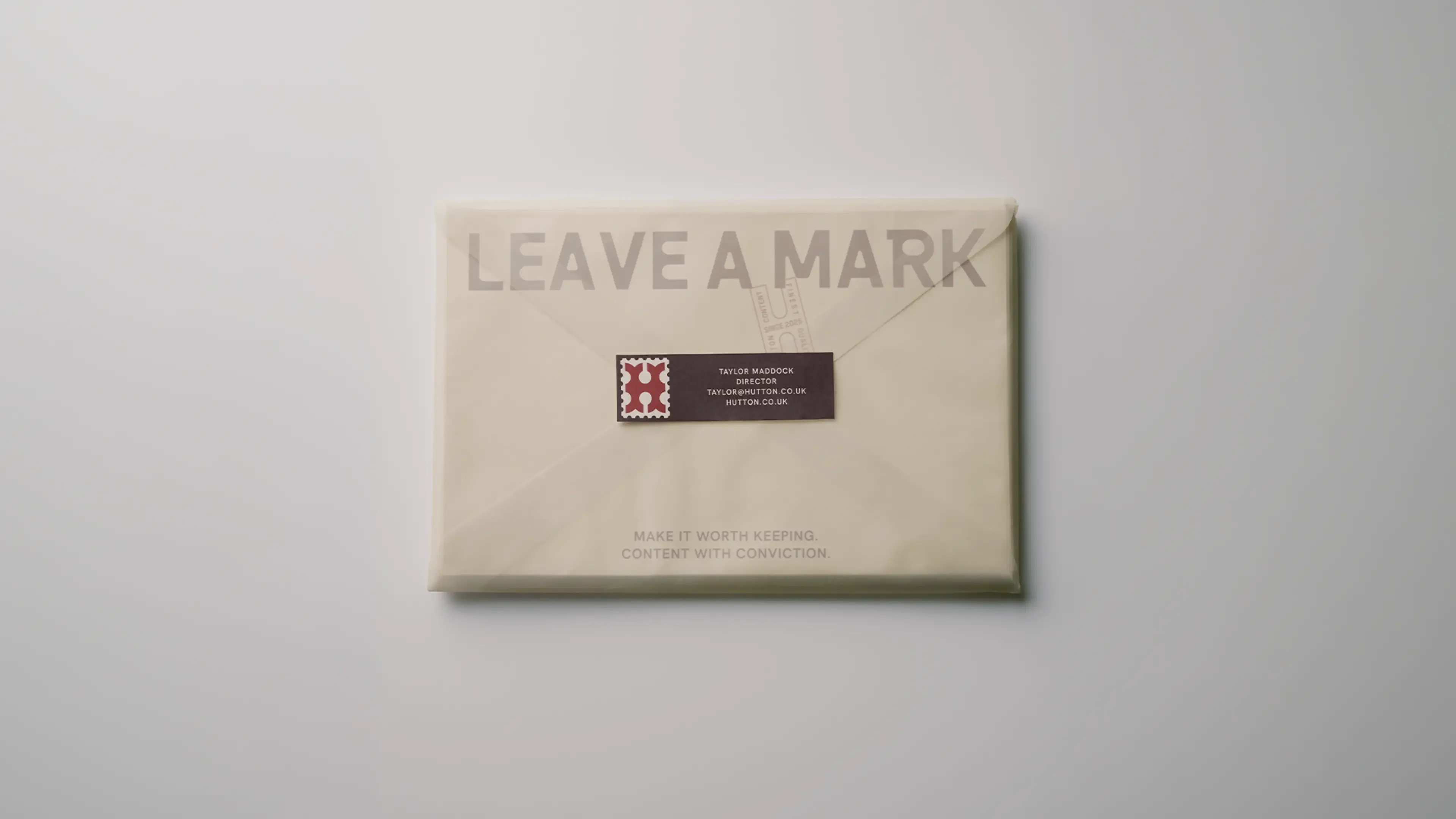 A cream envelope printed with “Leave a mark” in large capitals, sealed with a burgundy address label carrying a red monogram stamp. Along the bottom: “Make it worth keeping. Content with conviction.”