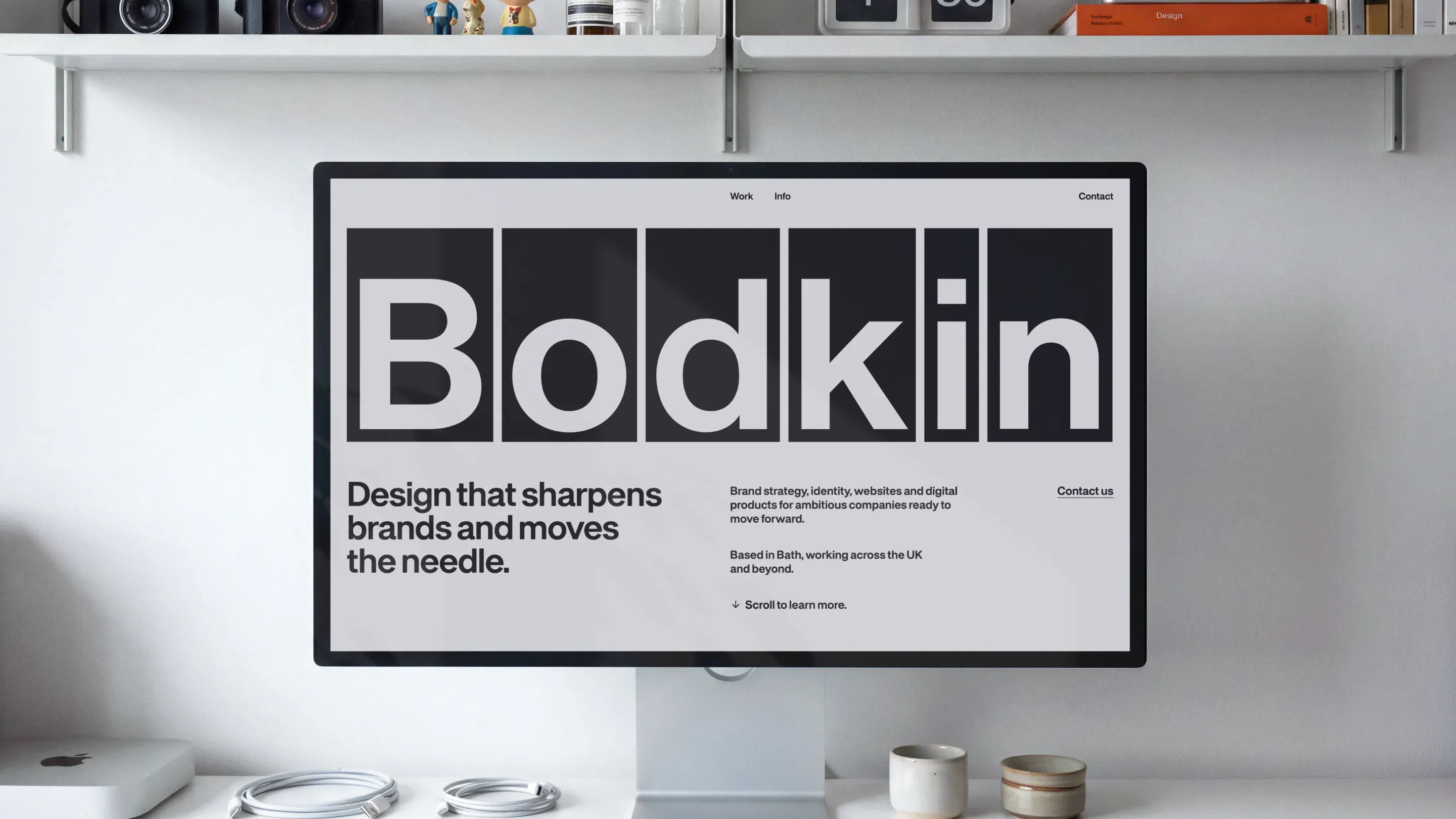 A computer monitor on a desk displays the Bodkin design studio website with the word Bodkin in bold letters and the tagline Design that sharpens brands and moves the needle, with shelves and decor in the background