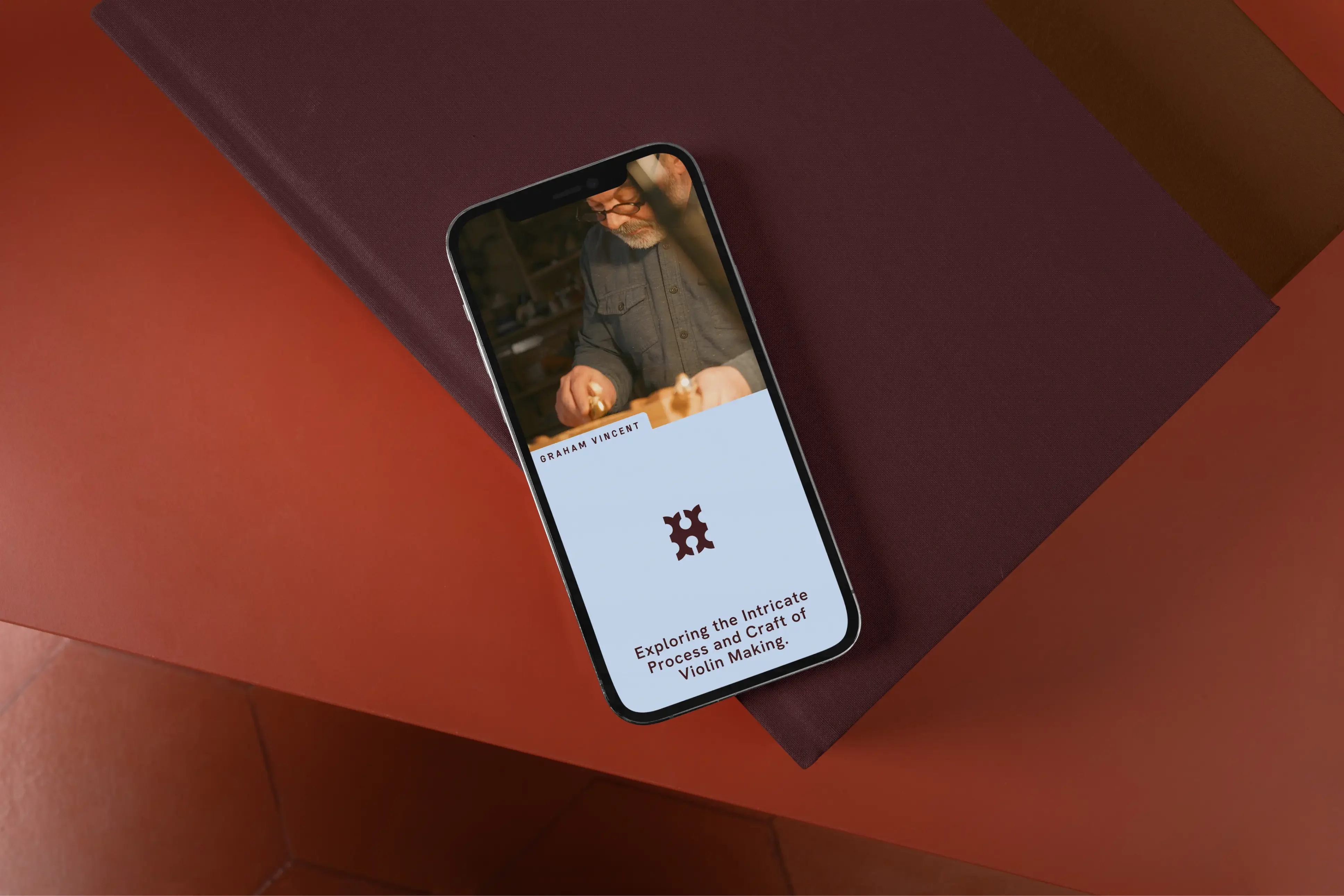 A phone resting on a burgundy cloth-bound book, showing a Hutton film about violin maker Graham Vincent with the monogram on pale blue