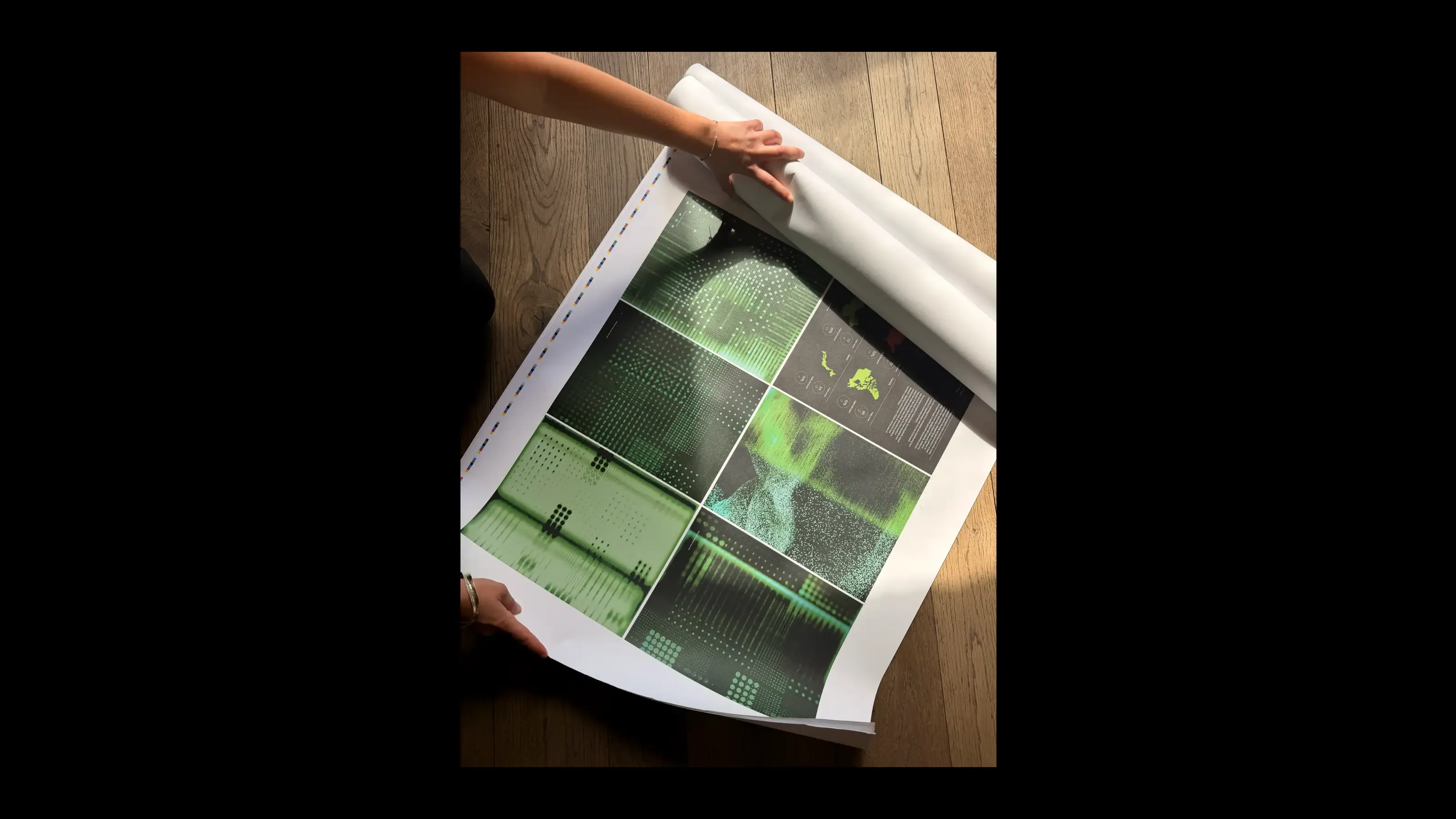 Two hands unrolling a large-format printed proof on a wooden floor, revealing a grid of green data-visualisation images: scan lines, particle fields, and halftone patterns being checked at full scale.