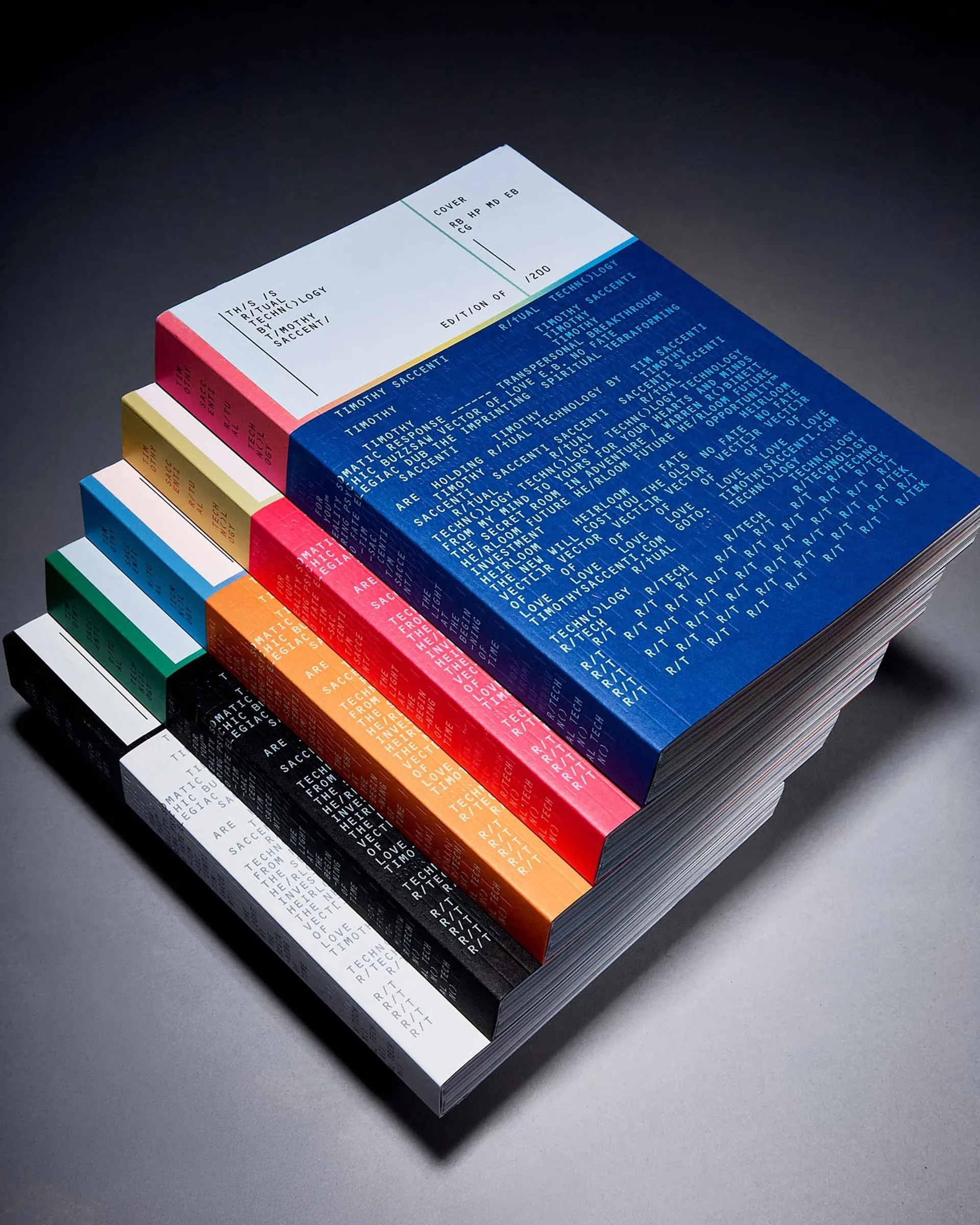 Six copies of the book Ritual Technology by Timothy Saccenti stacked in a staggered, fanned pile against a grey studio background. Each copy has a different cover and spine colour, deep blue on top, then pink, orange, black, and white beneath, and every cover, spine, and page edge is densely printed with columns of monospaced coded text, including the edition marking "edition of 200".